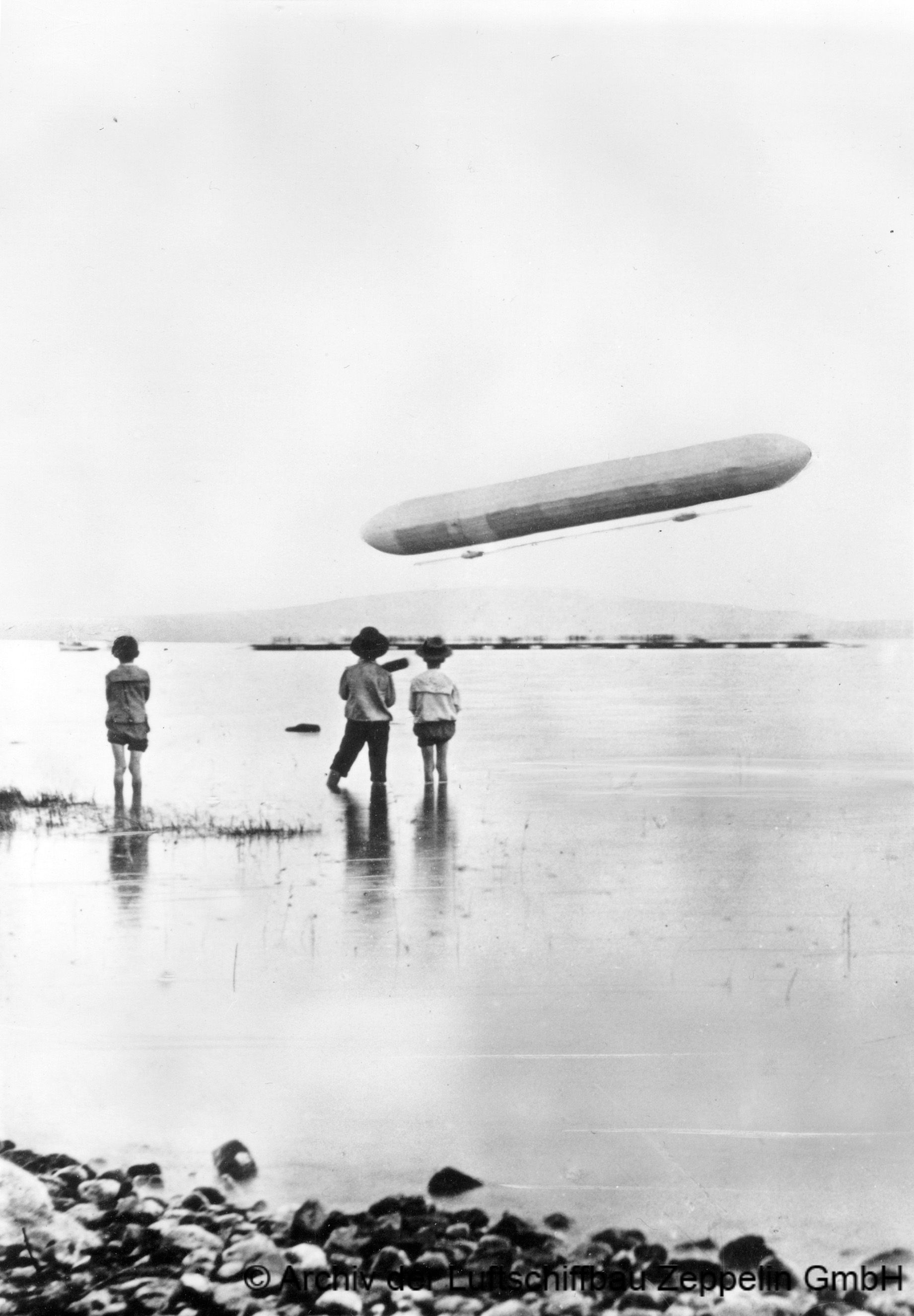 Drei Kinder beobachten vom Ufer des Bodensees den Aufstieg des Zeppelinschen Luftschiffs